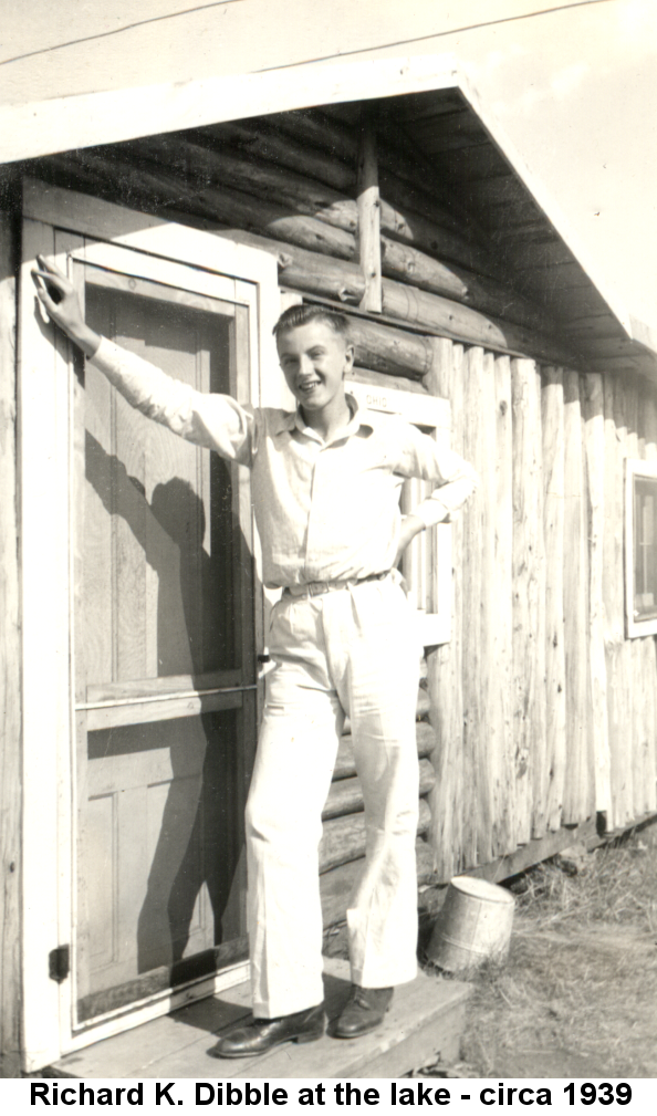 Black and white photo of teen-aged Richard K. Dibble in white long-sleeved shirt, white pants and black shoes standing on the wooden stoop or a log cabin, right arm extended and leaning against the closed screen door. An upside-down metal bucket is on the ground behind him.